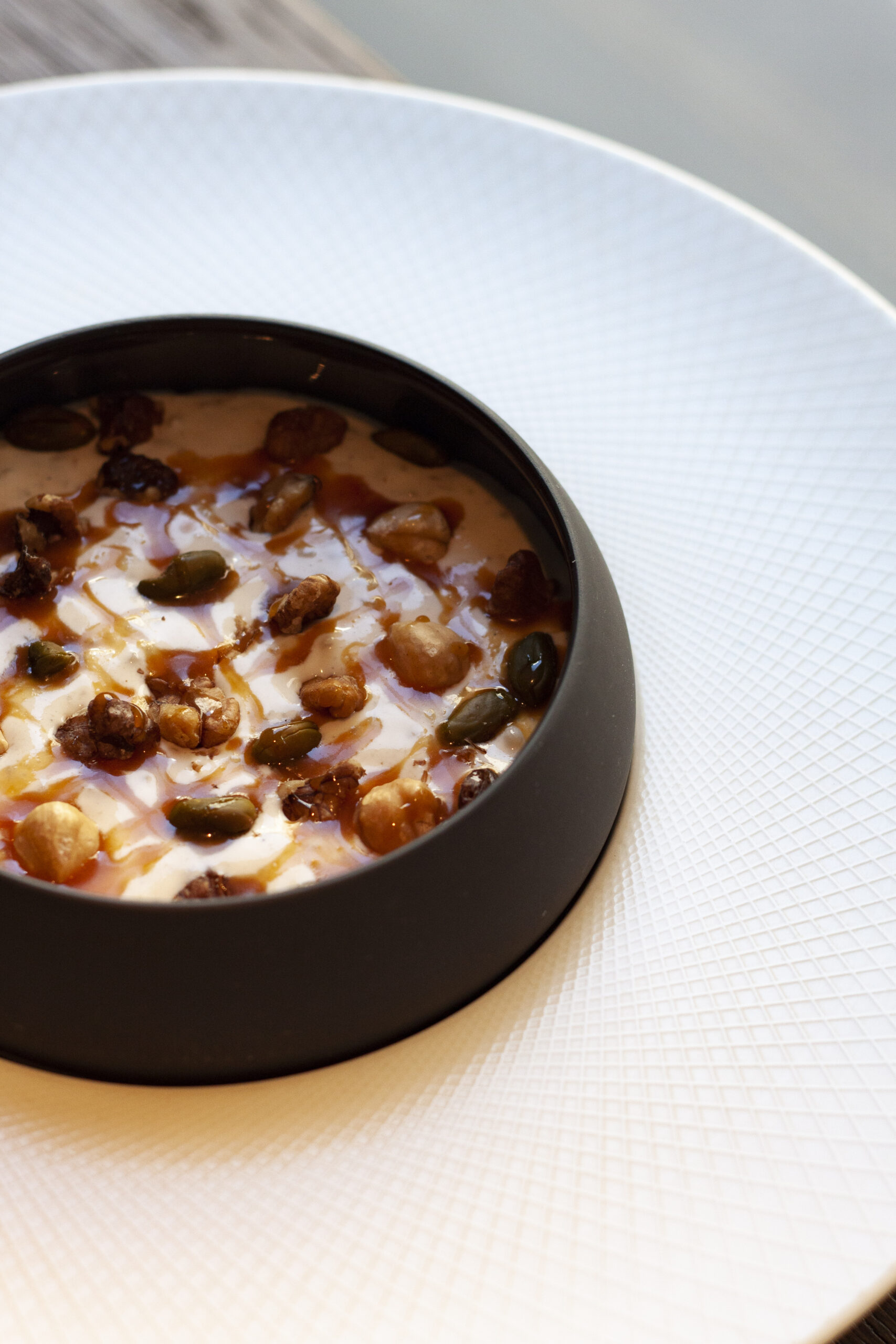 Dessert culinaire artisanal servi par un artisan pâtissier, garni de fruits secs croquants et d’un filet de caramel dans un bol noir sur une assiette blanche texturée