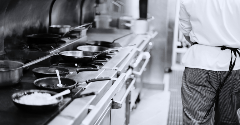 Chef vu de dos devant une rangée de poêles en cuisine professionnelle - Unsplash